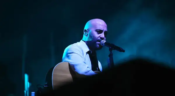 Giuliano Sangiorgi dei Negramaro, con la sua chitarra, canta al microfono, un passo indietro sul palco illuminato da luci blu.