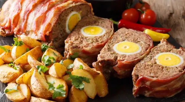 Pyszny klops z jajkiem w środku, zawinięty w boczek, podany z pieczonymi ziemniakami i warzywami.