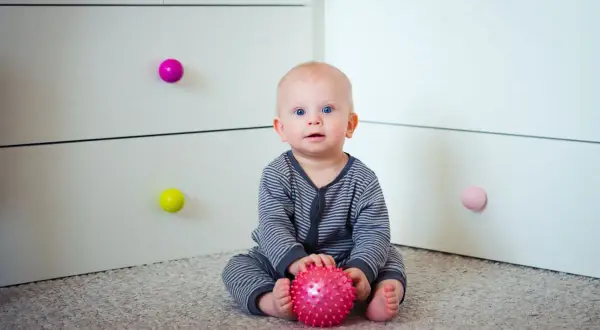 7 miesięczne dziecko z niebieskimi oczami siedzi na dywanie i bawi się różową, kolczastą piłeczką.