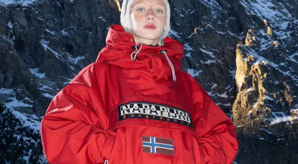 Modelka w czerwonej kurtce z logo Napapijri Geographic i flagą Norwegii na tle ośnieżonych gór. Napapijri co to za firma? To marka odzieży outdoorowej.
