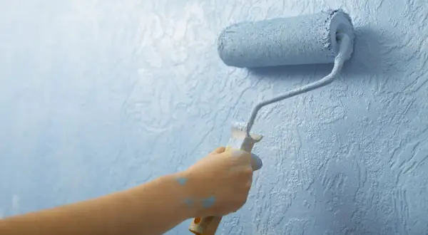 Mano pintando una pared con rodillo azul. El plástico del rodillo se ve salpicado de pintura.