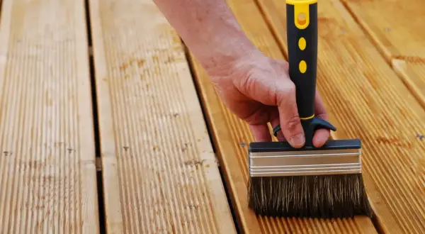 Main d'une personne utilisant une large brosse pour nettoyer bois après ponçage. La terrasse en bois est prête pour le traitement.