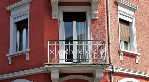 Dettaglio di un muro esterno con finitura a rasatura, finestre con persiane e un piccolo balcone con ringhiera in ferro battuto.