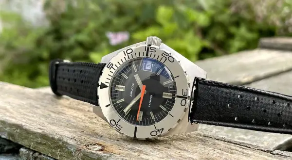 Reloj Sandoz Geneve Typhoon con correa negra, sobre madera rústica y fondo verde.