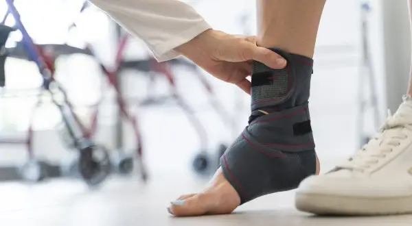 Dłoń zakłada ściągacz na kostkę, by zapewnić jej stabilizację. W tle widać rehabilitacyjny sprzęt.