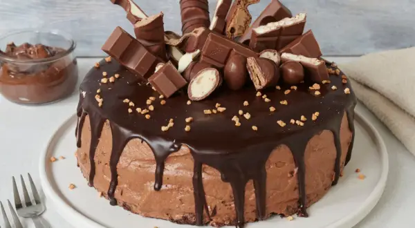 Eine üppige Nutella Torte, dekoriert mit Schokoriegeln und Nüssen. Daneben ein Glas Nutella.