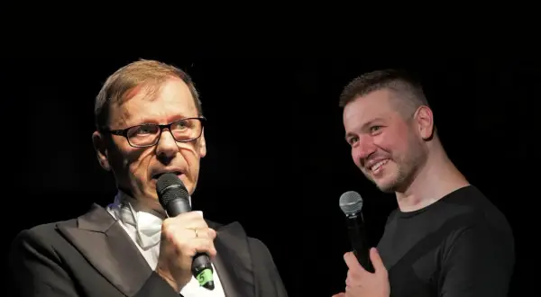 Dwóch mężczyzn z mikrofonami na scenie. Jeden w garniturze, drugi w czarnej koszulce. Widać, że socha stand up.