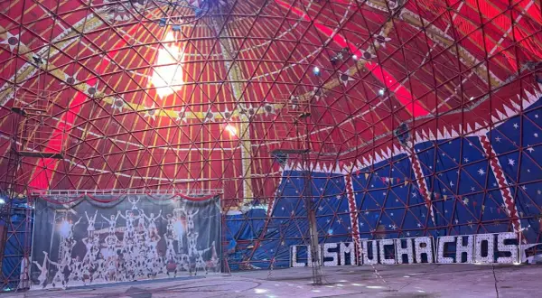 Interior de carpa de circo con techo rojo y azul, telón de fondo de acróbatas y luces que forman "MUCHACHOS". ¡Un espectáculo en Bempota, Ourense!