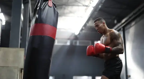 A boxer training intensely, sweat glistening on his skin as he throws punches at a heavy bag in a dimly lit gym.