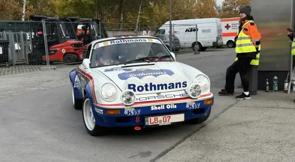 Ein weißer Porsche mit Rothmans-Aufschrift und dem Kennzeichen LB 07. Im Hintergrund stehen Gabelstapler und ein Rettungswagen.