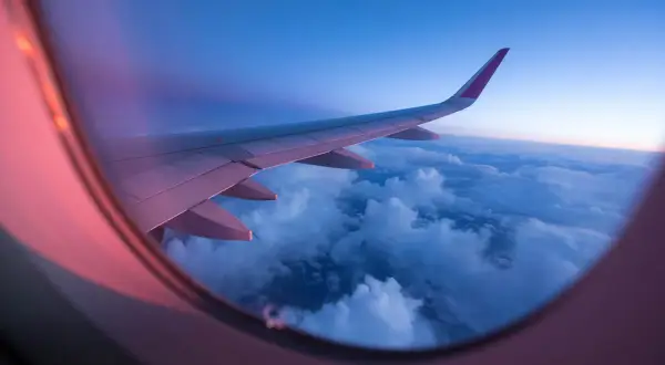 View from an airplane window at sunset. The wing stretches across the frame, bathed in pink and purple light, over a sea of clouds.