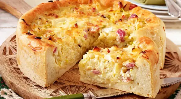 Ein goldbrauner Zwiebelkuchen mit Speck, ein köstliches zwiebelkuchen originalrezept, auf einem Holzbrett mit einem Messer.