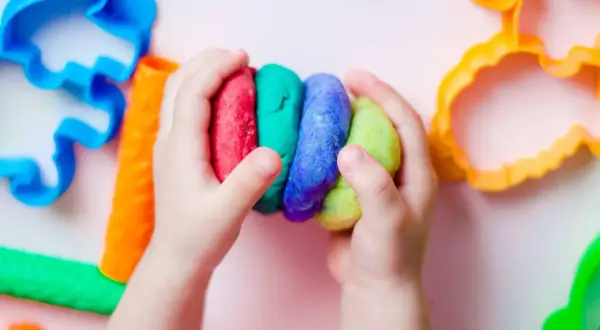 Kinderhände halten bunte Knetmasse, bereit zum Kneten. Nebenan liegen bunte Ausstecher, perfekt zum Knetseife selber machen.