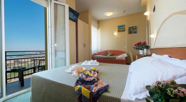 Pokój hotelowy z widokiem na morze, idealny na loty do Rimini. Na łóżku leży słomkowy kapelusz i kolorowy szal.