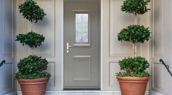 Une porte d'entrée grise avec une fenêtre décorative, encadrée par deux grands pots de plantes taillées. Idéal pour savoir comment changer une porte d'entrée.
