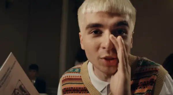 Młody mężczyzna z blond włosami, w kamizelce w kolorowe wzory, trzyma w ręku kartkę z tekstem, jakby czytał papuga.
