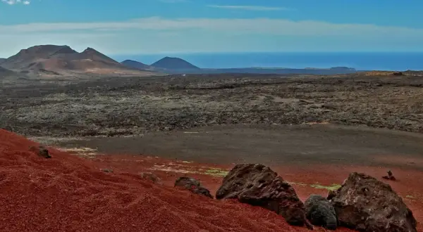 Krajobraz w parku narodowym Timanfaya: czerwone wzgórza, pola lawy i góry na tle oceanu.