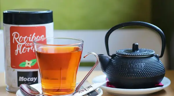 Szklanka z bursztynowym naparem, obok czajniczek i puszka z napisem "Rooibos con Flor". Czas na chwilę relaksu z rooibos.