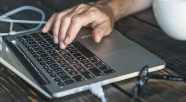 Dłoń na klawiaturze laptopa, biały kabel podłączony. Czas dowiedzieć się, jak zresetować laptopa.