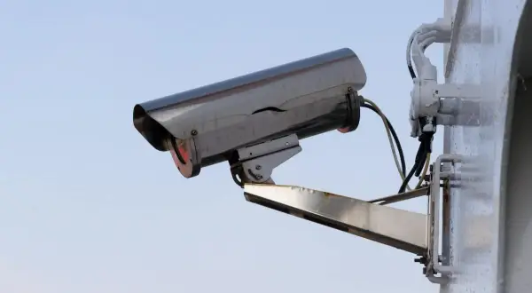 Caméra de surveillance extérieure, fixée sur un mur blanc. Le ciel bleu clair est visible derrière. On dirait qu'il y a un probleme camera de surveillance pas d'image.
