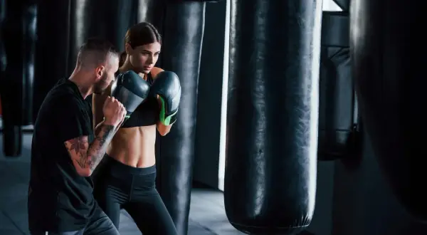 A man with tattoos trains a woman in boxing gloves. They are in a gym with punching bags, showcasing the physical and mental boxing benefits.