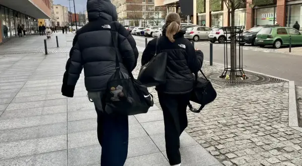 Dwie osoby w czarnych kurtkach puchowych idą chodnikiem, niosąc torby. Proste, miejskie kurtka puchowa stylizacje.