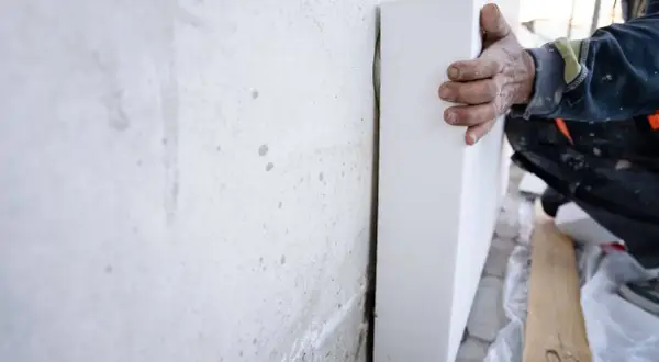Mano di un operaio che posiziona pannelli di polistirolo, eccellenti materiali isolanti, contro un muro grezzo per l'isolamento termico.