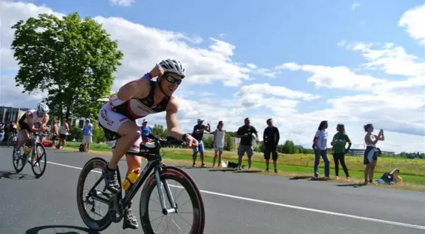 Triathlonista w kasku i okularach ściga się na rowerze, a widzowie dopingują go na poboczu. To część kalendarza triathlonowego.