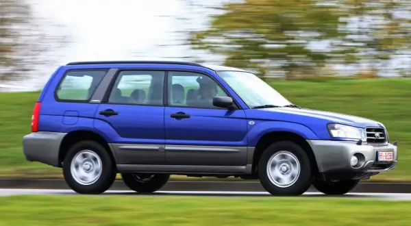 Niebieskie auto 4x4 Subaru Forester z napędem na cztery koła pędzi po drodze, otoczone zielenią.