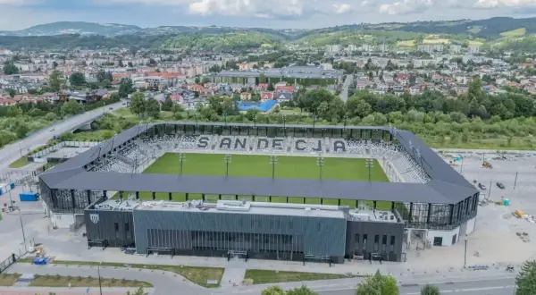 Nowoczesny stadion Sandecji z zieloną murawą i trybunami, otoczony miastem i wzgórzami.