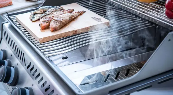 Fisch auf einem Holzbrett auf einem Grill, der zeigt, was ein Smoker Grill kann: Rauch steigt auf und veredelt das Gargut.