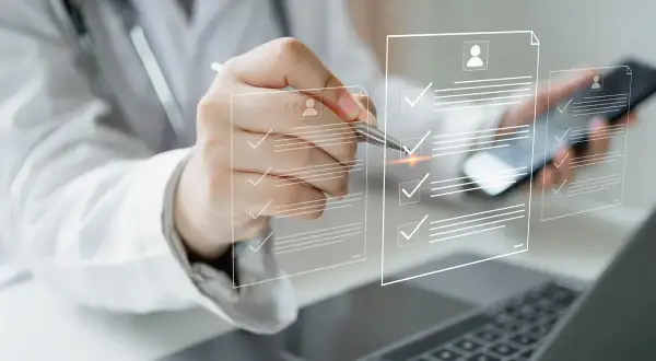Lekarz w białym kitlu z długopisem wskazuje na wirtualne dokumenty z listą zadań. W tle widać telefon i laptop. To może być proces wystawiania zwolnienia lekarskiego od psychiatry płatnego 100%.