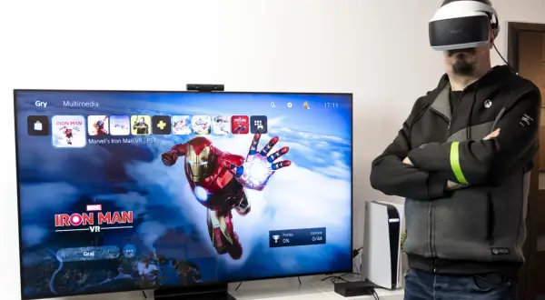 Mężczyzna w goglach VR obok telewizora z grą Iron Man VR.