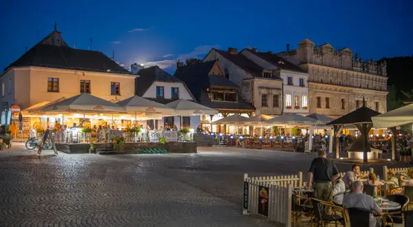 Wieczorny rynek w Kazimierzu Dolnym tętni życiem. Ludzie siedzą przy stolikach pod parasolami, ciesząc się letnim wieczorem.