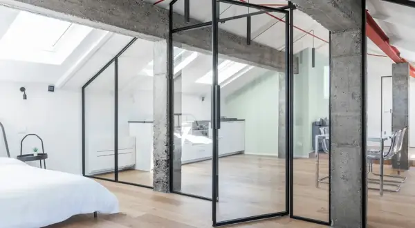 Moderno recupero sottotetto con pareti in vetro, travi a vista in cemento e parquet chiaro. Ambiente luminoso con cucina e zona living.