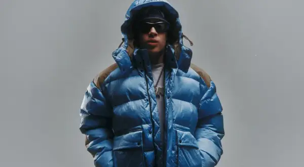 Giovane con piumino blu acceso, cappuccio alzato, occhiali da sole e una collana con ciondolo. Lo sfondo è un grigio chiaro, quasi low red.