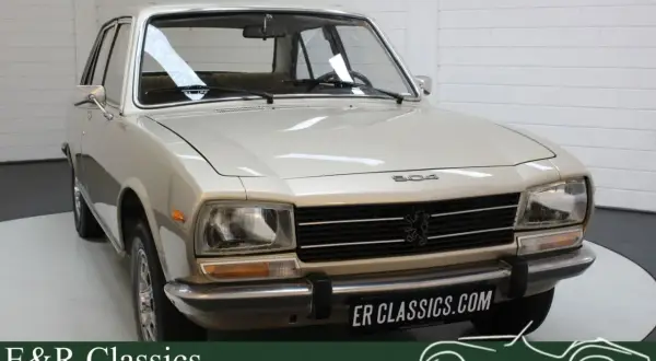 En ljusbeige Peugeot 504 står i ett garage. Bilen har en klassisk design med kromade detaljer.