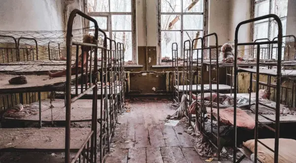 Opuszczony pokój z rzędami zardzewiałych łóżek piętrowych, zniszczonymi oknami i śmieciami na podłodze.