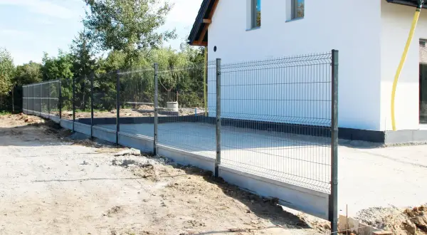 Trwa budowa ogrodzenia wokół nowoczesnego domu. Metalowa siatka na betonowym fundamencie otacza posesję.