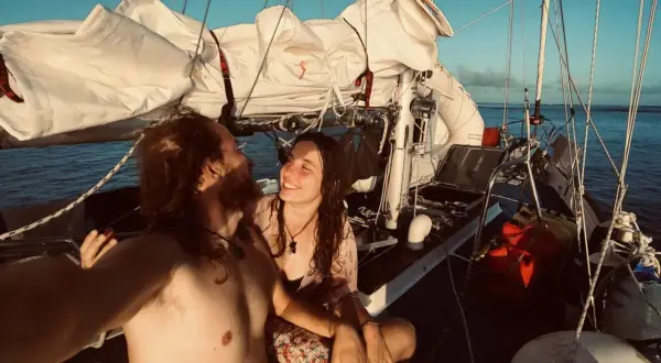 Coppia felice in barca a vela al tramonto, con vele bianche spiegate e mare calmo.