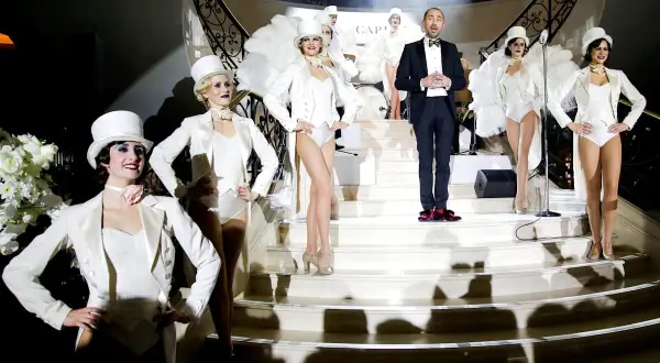Bühnenauftritt mit Tänzerinnen in weißen Outfits und einem Mann im schwarzen Anzug mit **white tie**.