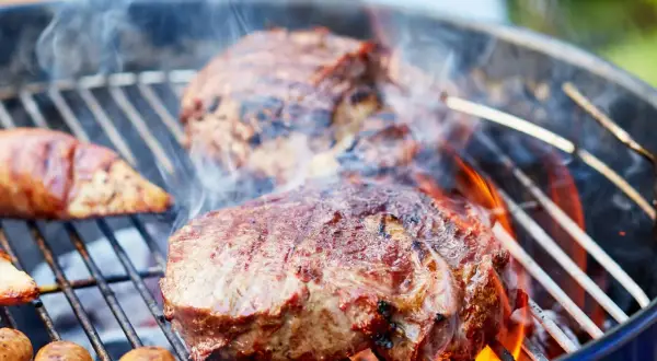 Saftiges Fleisch zum Grillen zischt auf dem Rost. Würstchen und Steaks brutzeln über heißen Kohlen, umgeben von Rauch und Flammen.
