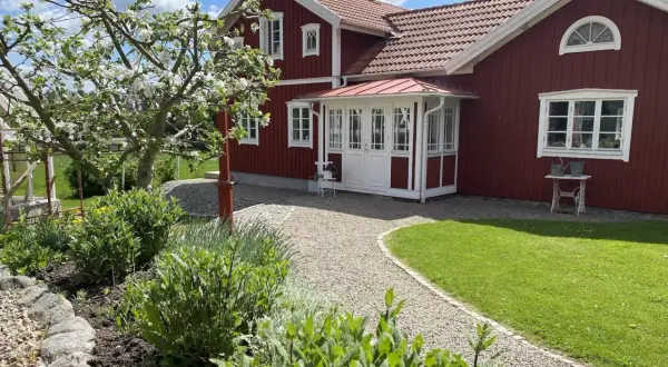 Ett rött hus med vita detaljer och en vacker farstukvist som leder in till hallen. En blommande äppelträd och grönskande trädgård.