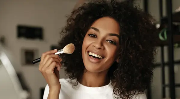 Donna sorridente con capelli ricci applica cipria con un pennello, mostrando come mettere la cipria senza pennello.