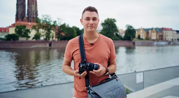 Mężczyzna z aparatem w rękach, w plenerze nad rzeką, z widokiem na zabytkowe budynki i katedrę.