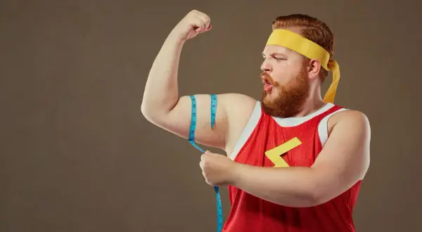 Mężczyzna typu endomorfik z wąsami i brodą, w żółtej opasce i czerwonej koszulce, mierzy obwód bicepsa.