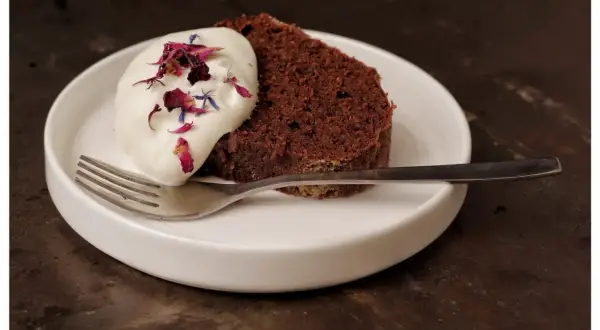 En bit saftig sockerkaka choklad med vispgrädde och blomblad på en vit tallrik med en gaffel.
