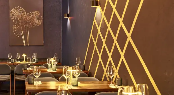 Stilvolles Ambiente im Lovis Restaurant: gedeckte Tische mit Gläsern und Kerzen, dunkle Wände mit goldenem Muster und ein Kunstwerk mit getrockneten Blumen.