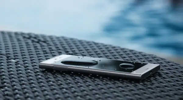 Smartfon z kroplami wody na ekranie, leżący na plecionej powierzchni. W tle widać basen. To dowód, że ip67 co oznacza - ochronę przed wodą.