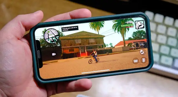 Ręka trzyma smartfon z grą RPG na telefon. Na ekranie widać postać na rowerze przed domem, palmy i ikony sterowania.